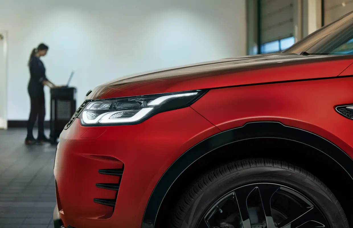 The front of a red Land Rover Discovery Sport in a service bay with a technician at a diagnostic station behind